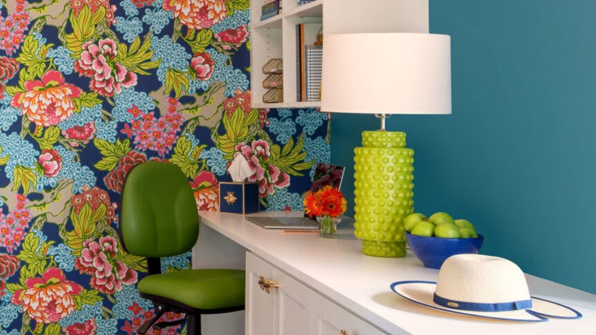 Close-up of a white desk styled with the Robert Abbey Dolly Table Lamp in Citron glaze, a green task chair, straw hat, and bowl of green apples against floral wallpaper and a teal wall.